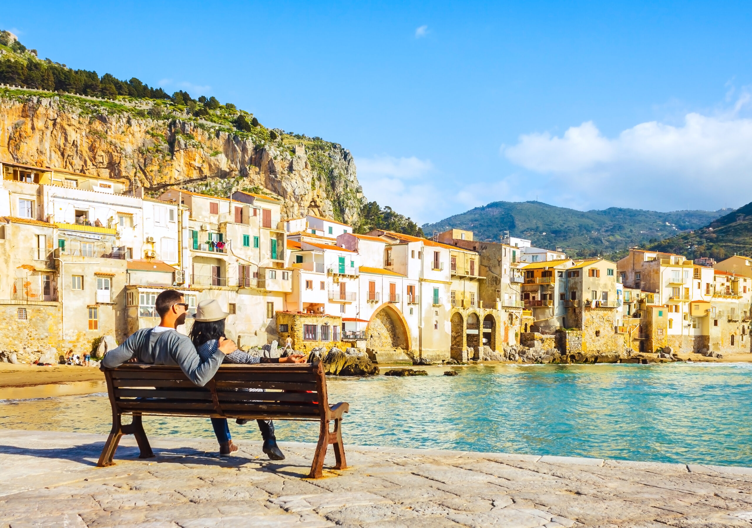Couple sitting on bench, enjoying view of beach town of Cefalu i